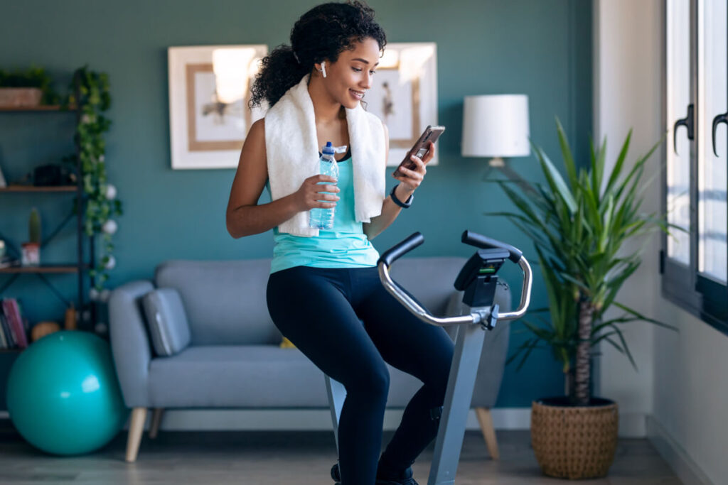 Afro young fitness girl using mobile phone while training on exercise bike at home to prevent spider veins